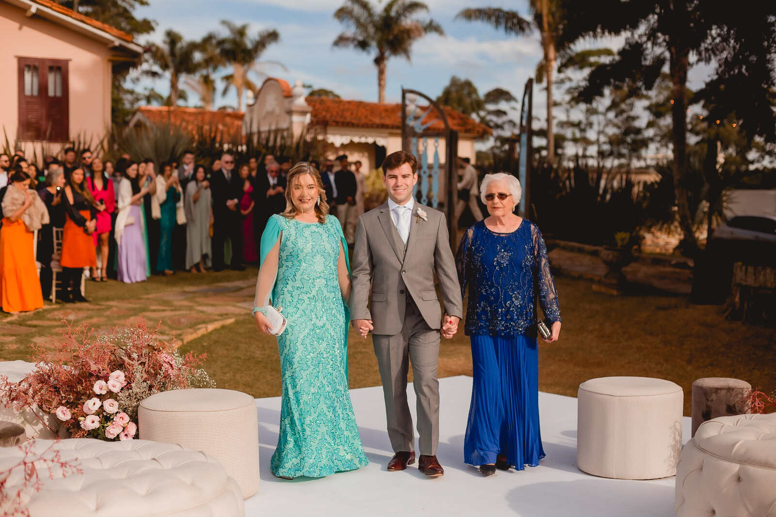 entrada do noivo com terno cinza ao lado da mãe com vestido azul tiffany e avó com vestido azul cobalto