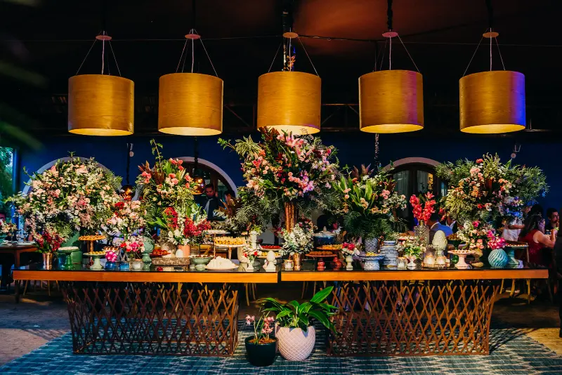 mesa de doces e bolo decorada com iluminação com luminárias pendentes cilíndricas com acabamento dourado que cria um ambiente acolhedor.
