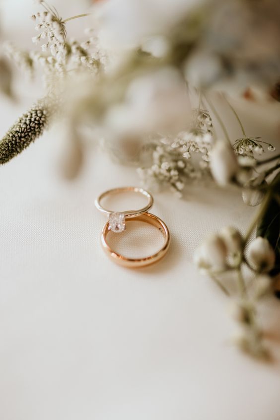 Ao centro da imagem estão uma aliança e um anel de noivado, ao redor algumas flores decoram a foto.