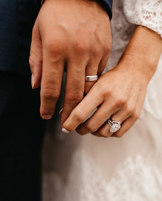 As mãos de um casal estão em foco, seus dedos se tocam. Na mão dele, no dedo anelar, está uma aliança e na mão dela, também no dedo anelar, um anel.