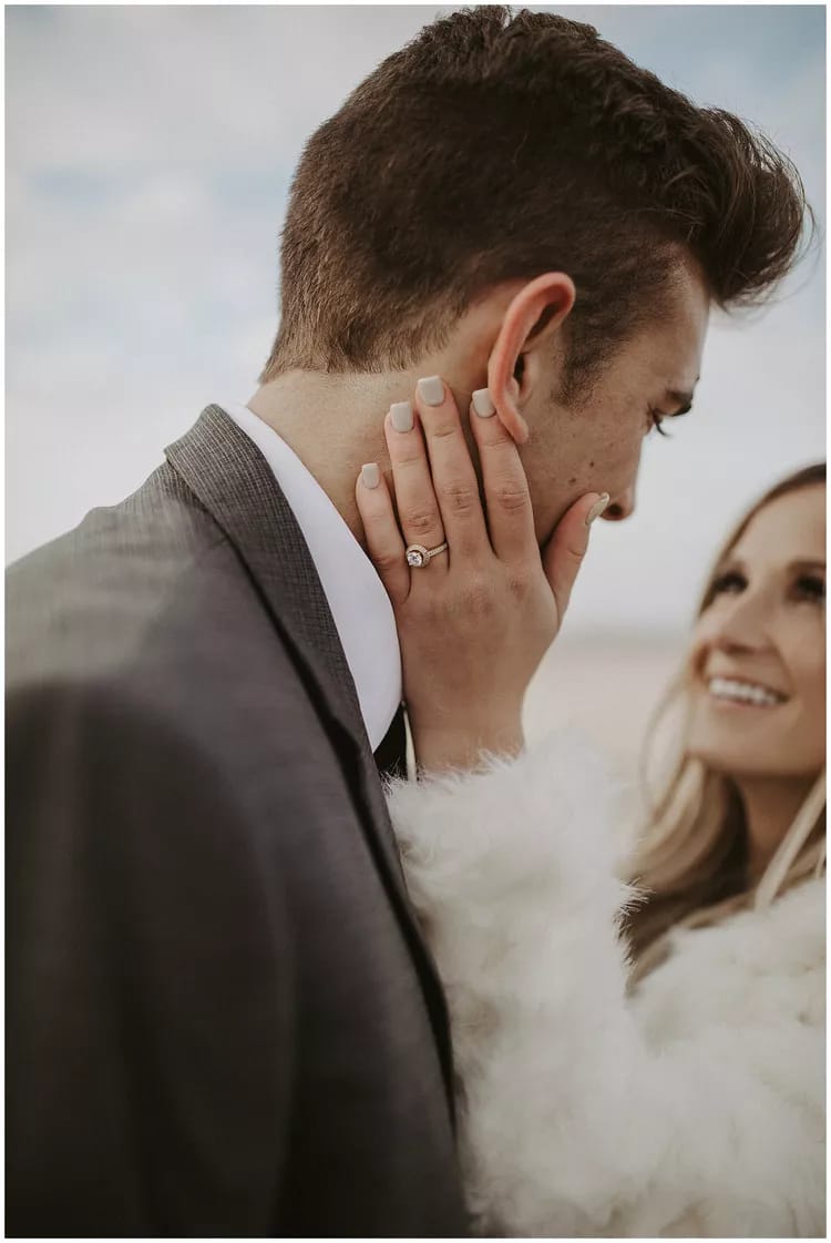 Um casal está sorrindo, o homem veste um terno preto e a mulher um casaco branco, ela está com sua mão no rosto dele e usa um anel de noivado.