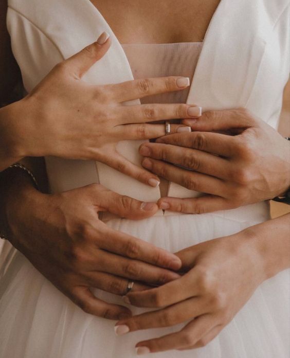 O tronco de uma noiva, de vestido branco, abraçada pelo noivo. Suas mãos estão em foco e eles trocam alianças douradas na mão esquerda.