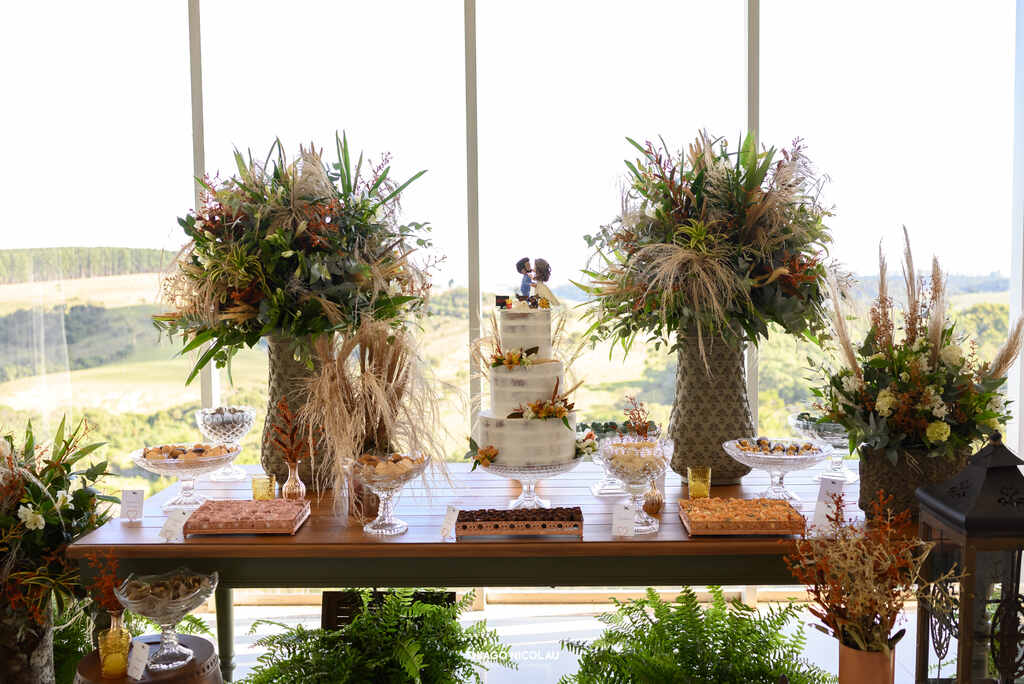 mesa com bolo de casamento branco semiespatulado com flores e topo de bolo com bonecos e ao ado vasos com plantas e folhas secas