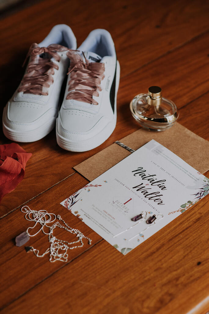 tênis de noiva branco com cadarço ros ao lado de frasco de perfume colar e convite de casamento