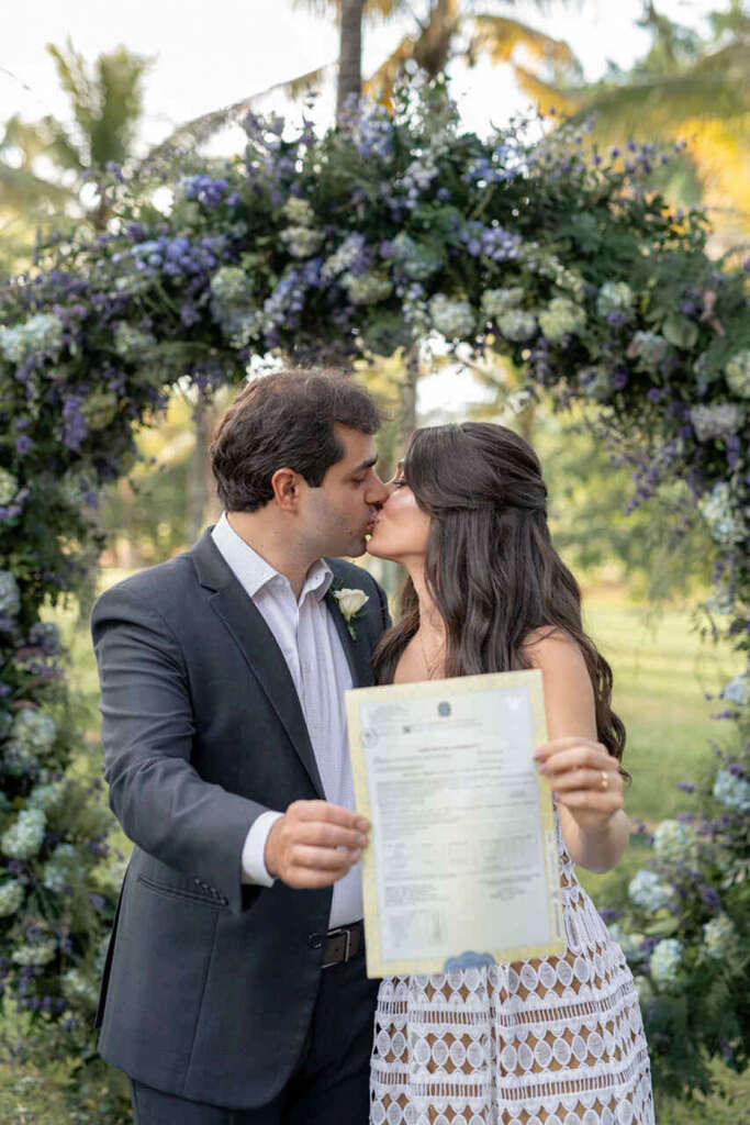 casal de noivos se beijando em frente a altar florido circular segurando certidão de casamento civil