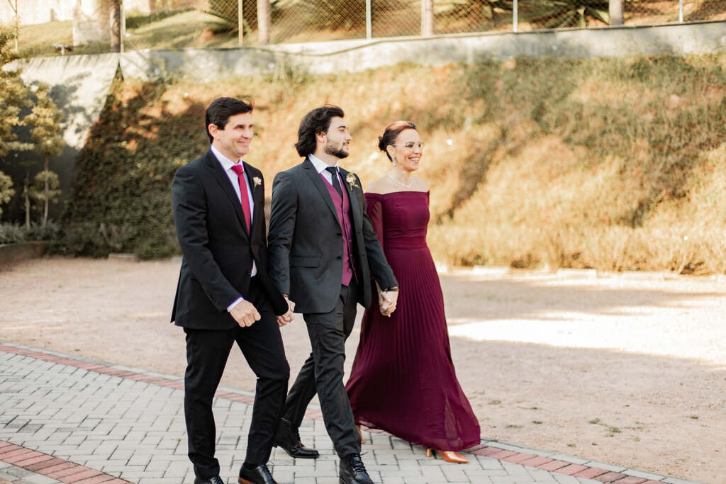 entrada do noivo com terno e gravata preta e colete na cor marsala ao lado do pai com tenro preto e gravata vermelha e mãe com vestido com mangas na cor marsala