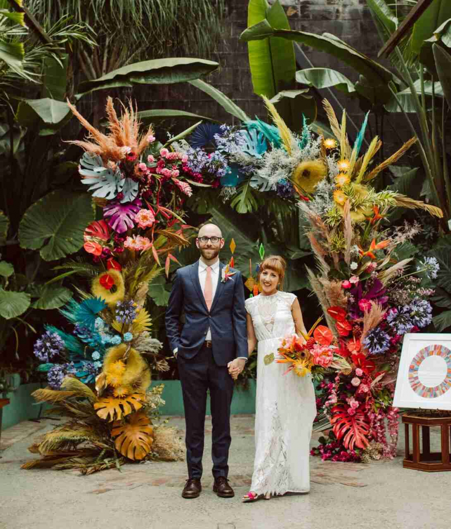 Casal de noivos em frente a um arco cheio de flores e folhagens coloridas