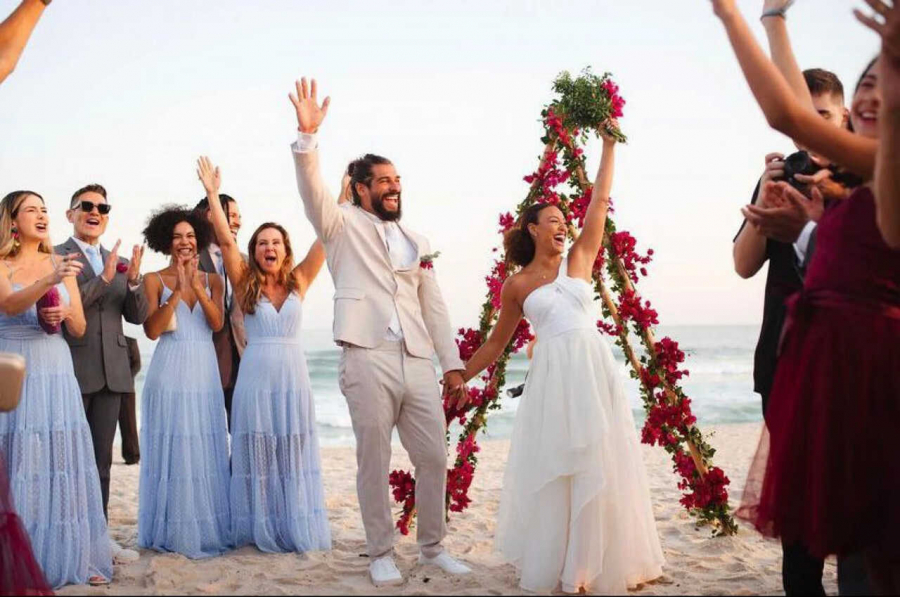 Casal com mãos levantadas comemorando casamento na praia, alguns convidados em volta