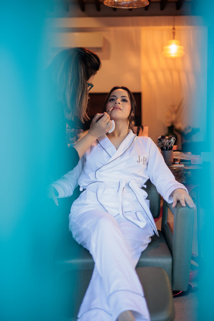 noiva de robe branco sentada na cadeira enquanto mulher faz sua maquiagem
