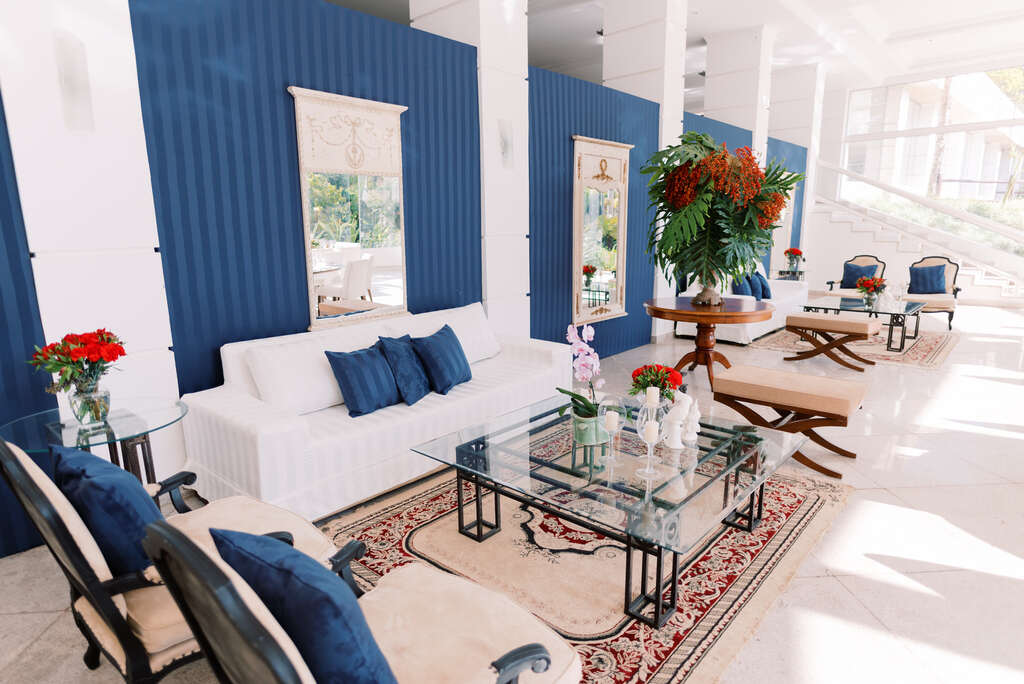 salão branco e azul clássico com lounge com sofá branco com almofadas azuis decorado com tapete e vasos com plantas e flores vermelhas