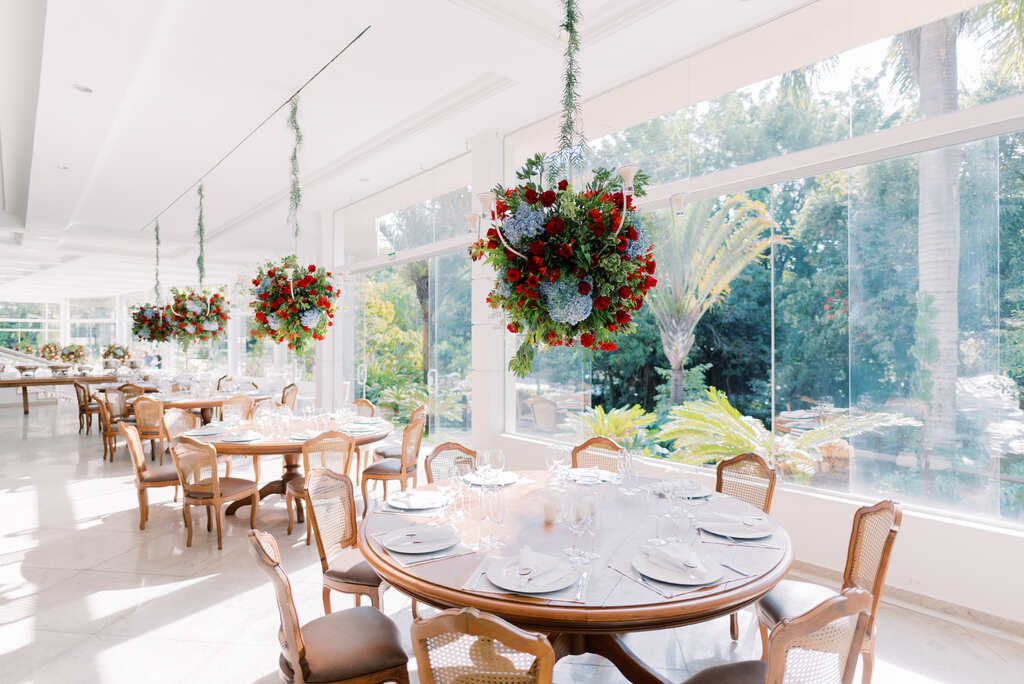 salão branco com mesas redondas postas decoradas com arranjos aéreos com rosas e hortensias azuis
