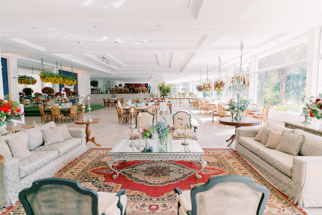 salão branco com lounge com sofá branco com almofadas azuis decorado com tapete e vasos com plantas e flores vermelhas