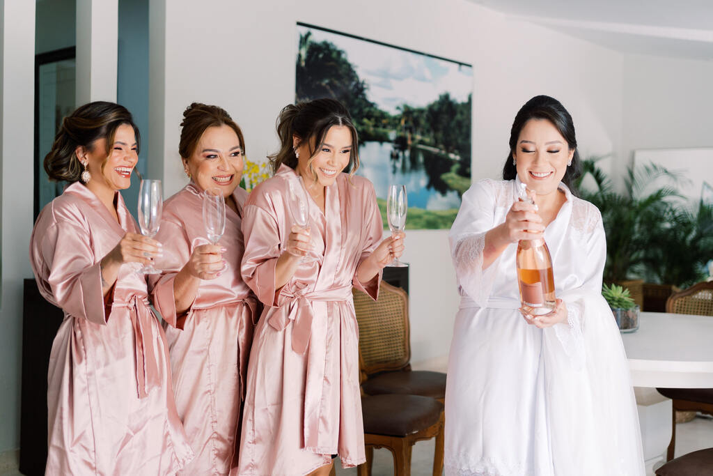 noiva com robe branco segurando bebida e ao lado três madrinhas com robes na cor rosê segurando taças