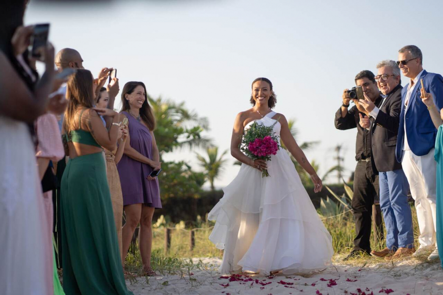 Noiva entrando sozinha na cerimônia na praia, com buquê de flores em tom magenta
