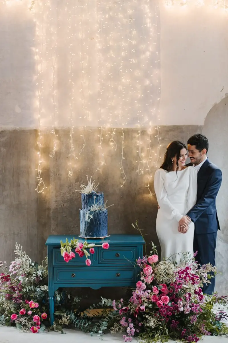 decora&ccedil;&atilde;o inclui um arranjo floral exuberante com tons de rosa e roxo na base da mesa. O fundo &eacute; composto por uma cortina de luzes quentes