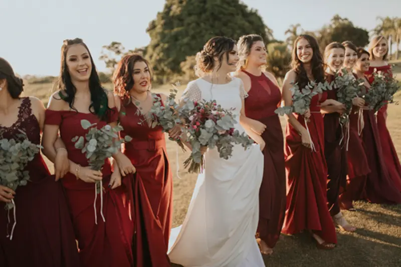 noiva caminhando no campo ao lado das madrinhas que est&atilde;o com vestido na cor marsala