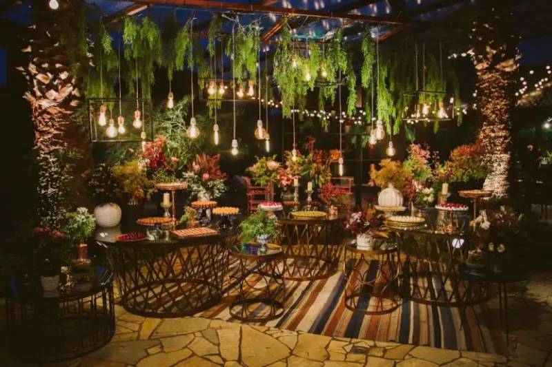 decora&ccedil;&atilde;o de recep&ccedil;&atilde;o de casamento com um estilo r&uacute;stico e intimista. O ambiente utiliza ilumina&ccedil;&atilde;o decorativa c&ecirc;nica, com diversas l&acirc;mpadas pendentes criando um efeito acolhedor sobre as mesas.