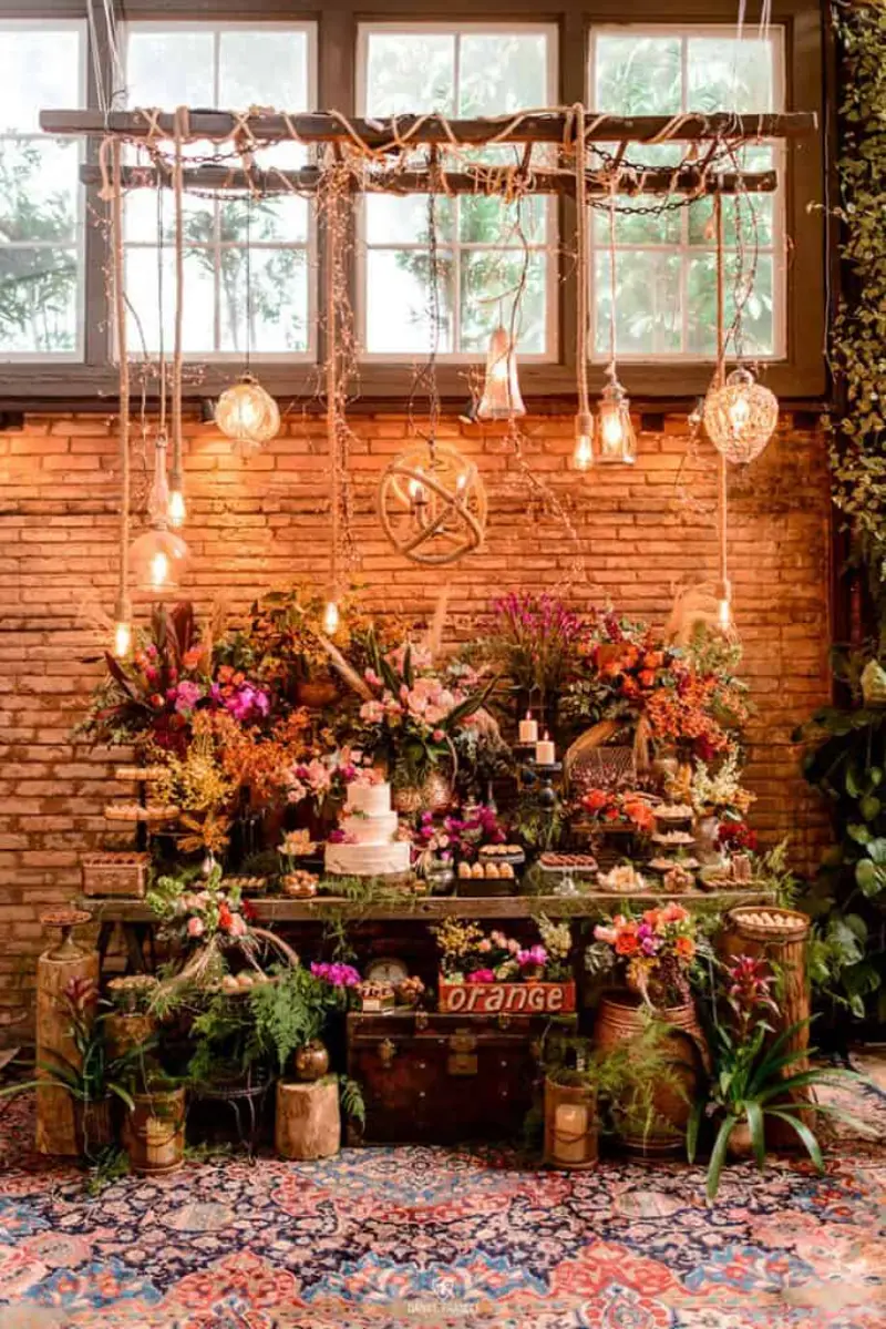 mesa de bolo e doces com parede de tijolos aparentes, ilumina&ccedil;&atilde;o com l&acirc;mpadas pendentes de estilo industrial/r&uacute;stico e arranjos florais densos.