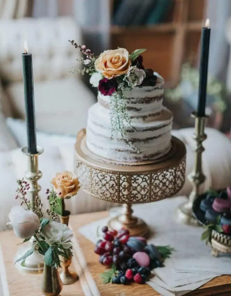 bolo de casamento estilo "naked cake" de dois andares, decorado com flores frescas em tons de pêssego e vinho, posicionado sobre um suporte de metal dourado