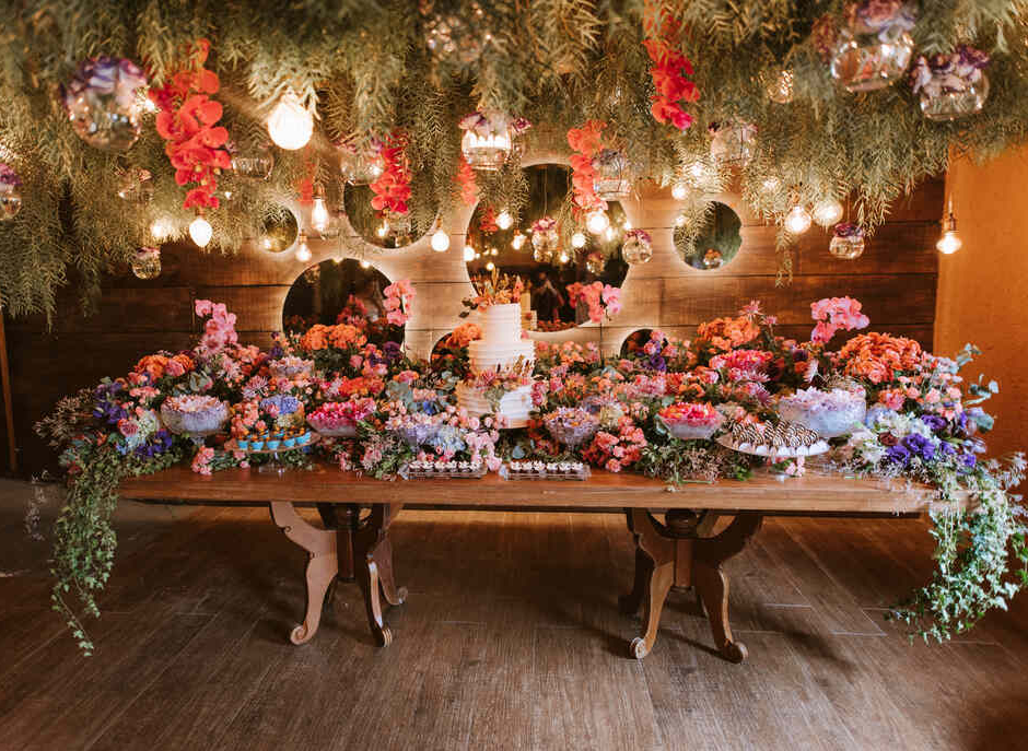 mesa com bolo de casamento com três andares e topo com flores e em volta muitas flores coloridas e bandejas com doces finos e em cima arranjos aéreos com plantas e orquideas