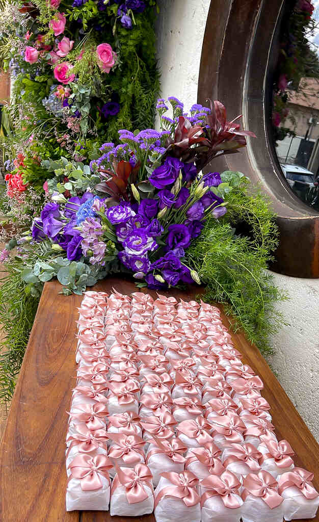 mesa com bem-casados com embalagem branca com lacinhos cor de rosa e ao lado arranjo com flores roxas
