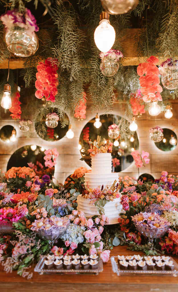 mesa com bolo de casamento com três andares e topo com flores e em volta muitas flores coloridas e bandejas com doces finos e em cima arranjos aéreos com plantas e orquideas e atrás da mes espelhos redondos iluminados