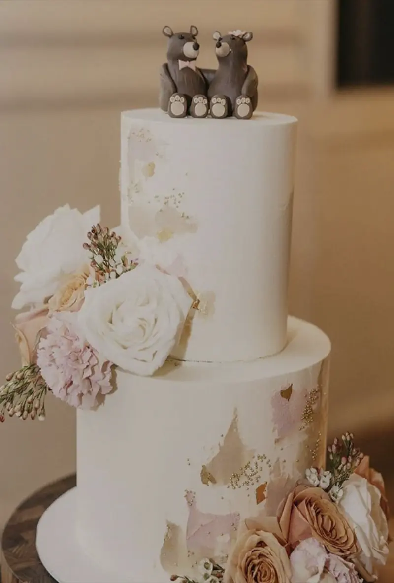 bolo de casamento de dois andares com cobertura branca lisa, decorado com flores frescas em tons de rosa e branco e um topo de bolo de ursinhos
