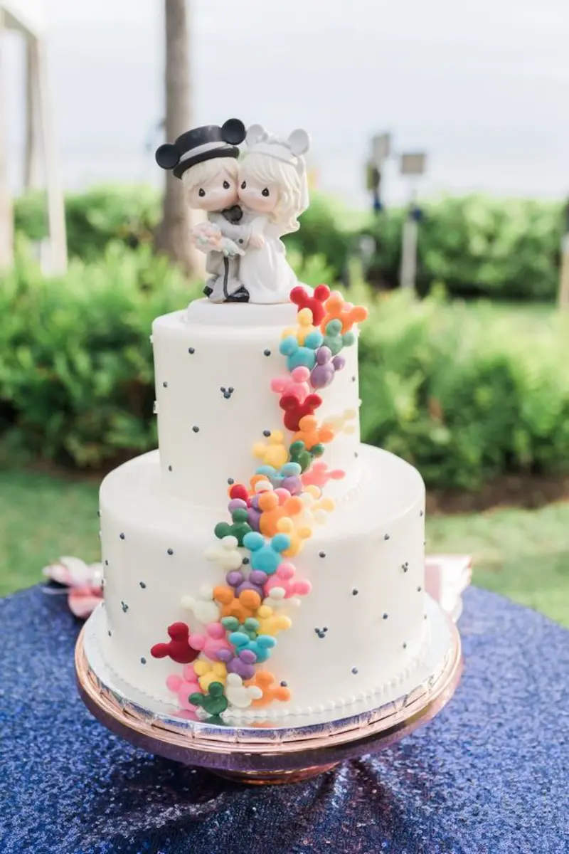 bolo de casamento inspirado no Mickey Mouse da Disney, apresentando um design de dois andares com detalhes coloridos de cabeças de Mickey e um topo de bolo romântico