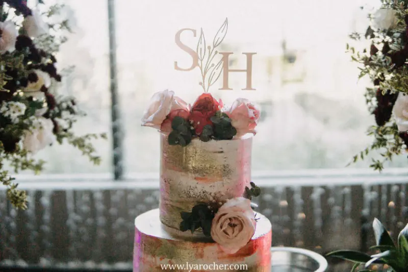bolo de casamento de estilo semi-naked com detalhes pintados em dourado, flores frescas em tons de rosa e vermelho, e um topo de bolo personalizado com as iniciais "SH
