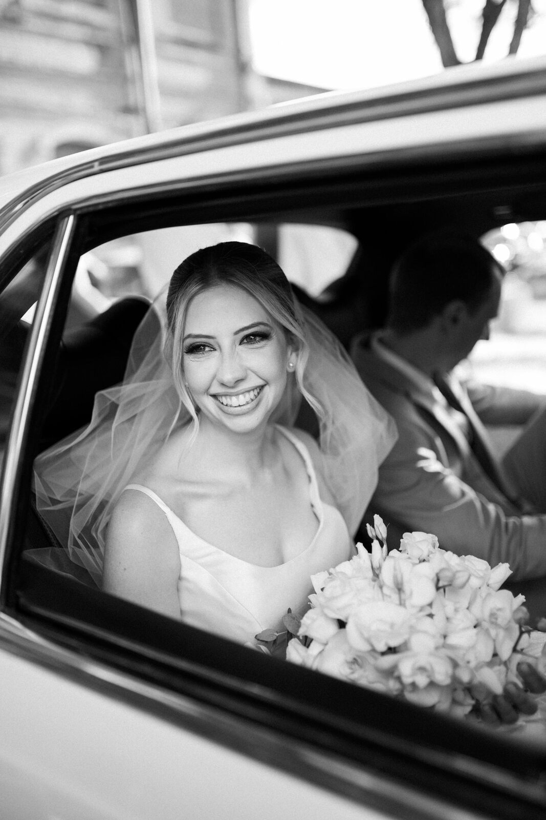 noiva com véu segurando buquê redondo com orquideas brancas sorrindo dentro do carro