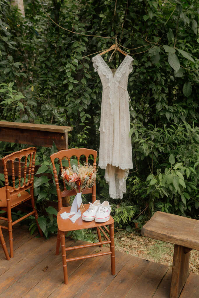 vestido de noiva boho com decote em v ao lado de cadeira de madeira com buquê com fita branca e tênis branco