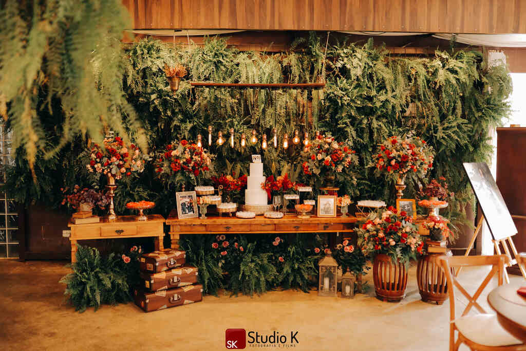mesa rústica com bolo de casamento branco ao lado de vaos com flores vermelhas