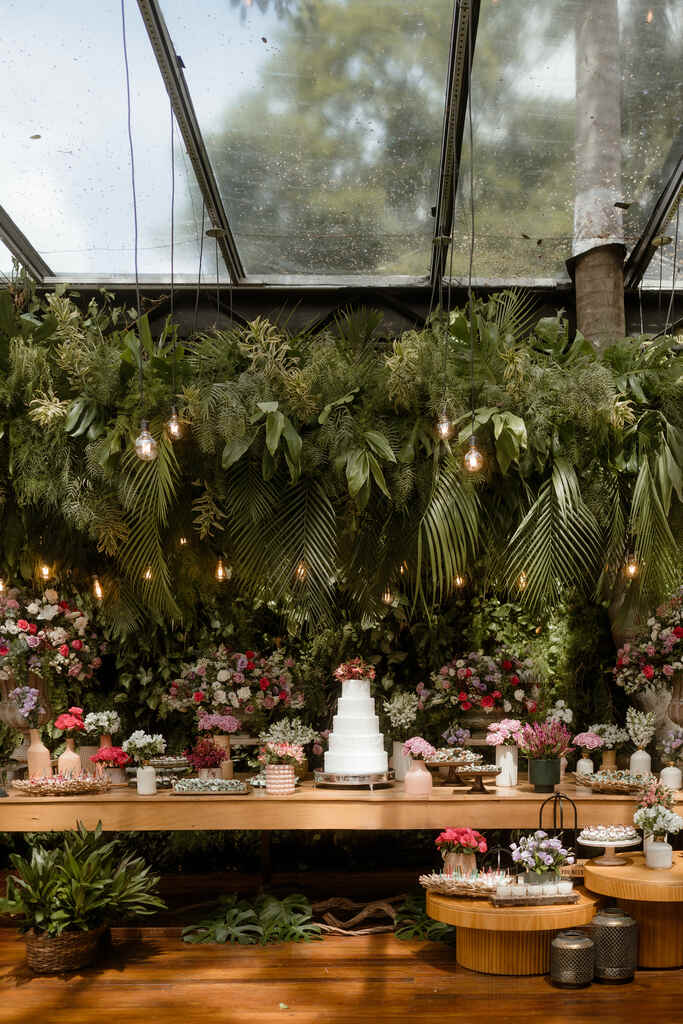 mesa com bolo de casamento branco com cinco andares com topo de flores e ao lado bandejas com doces de casamento e flores brancas e rosas