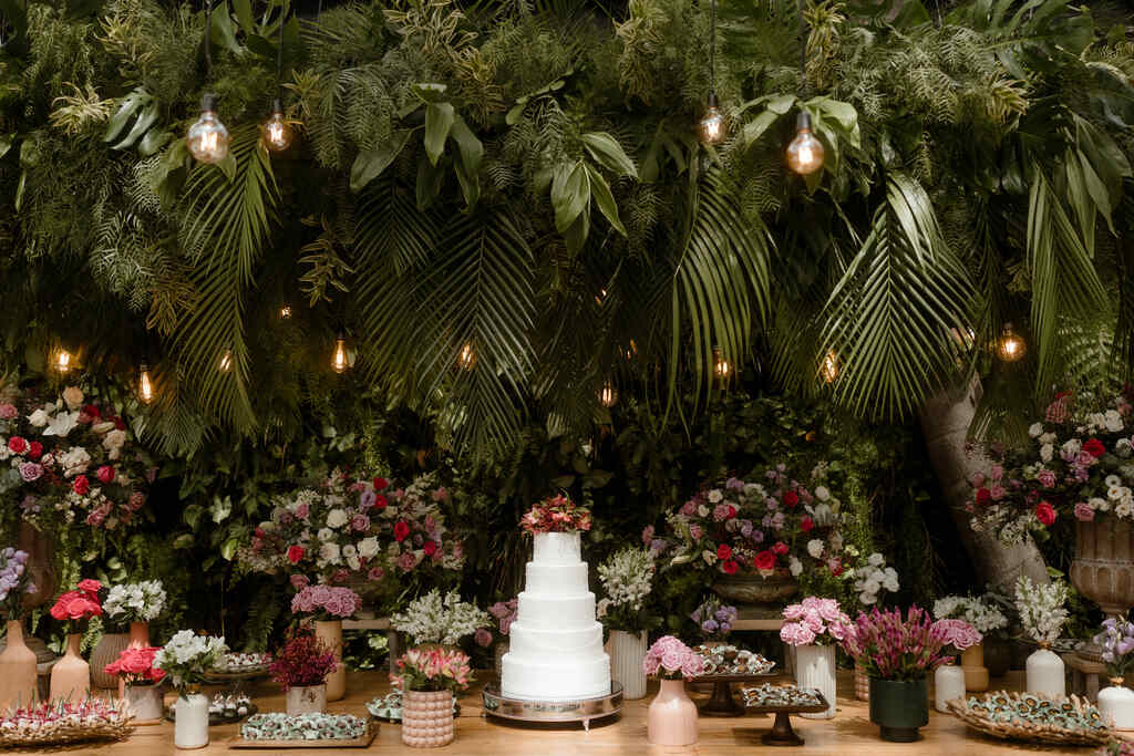 mesa com bolo de casamento branco com cinco andares com topo de flores e ao lado bandejas com doces de casamento e flores brancas e rosas