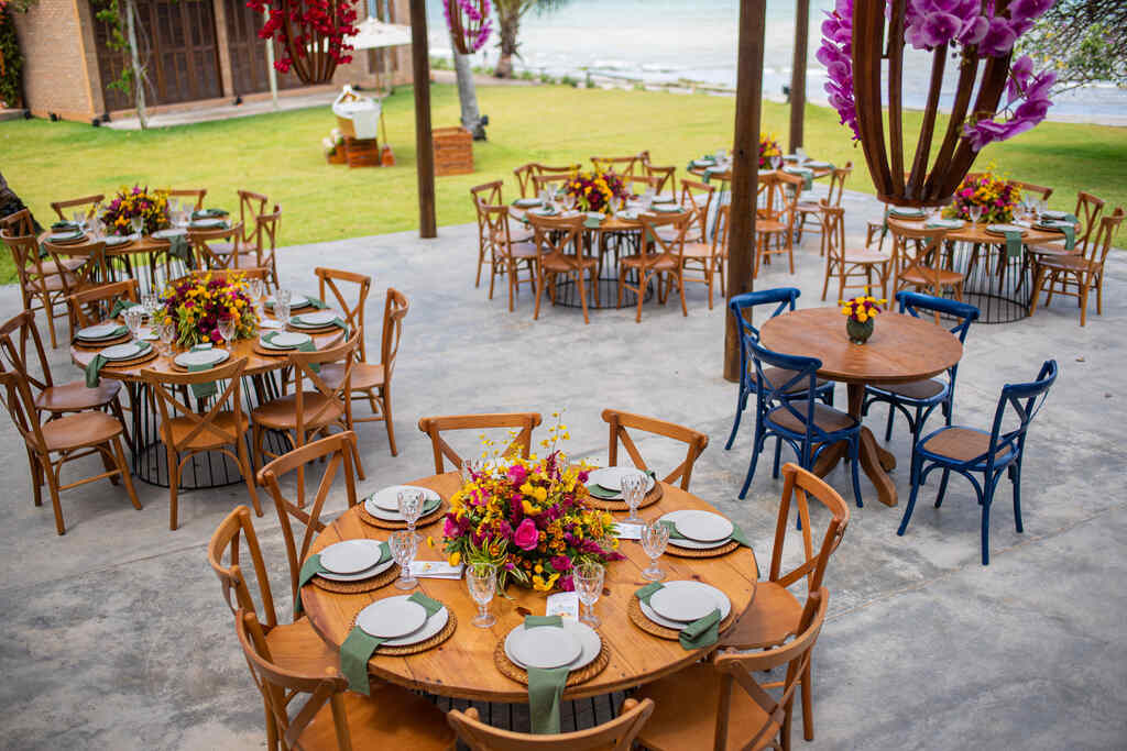 salão ao ar livre com mesas redondas postas com centro de mesa com flores rosas e amarelas