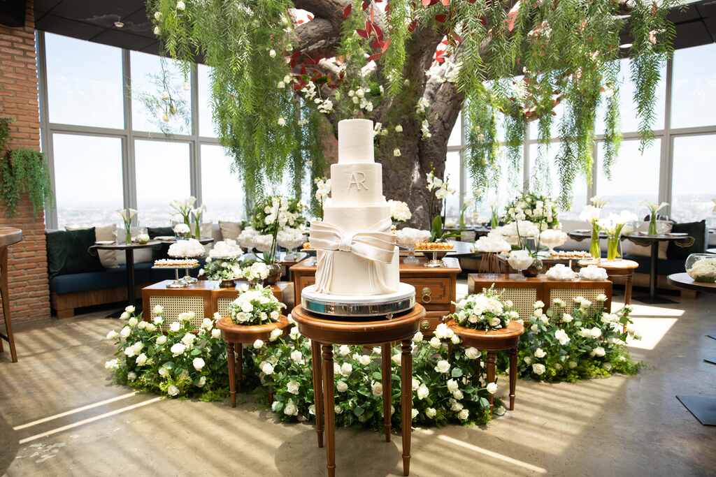 salão com árvore no centro com mesa decorada com ropsas brancas doces e na frente bolo de casamento com quatro andares com laço