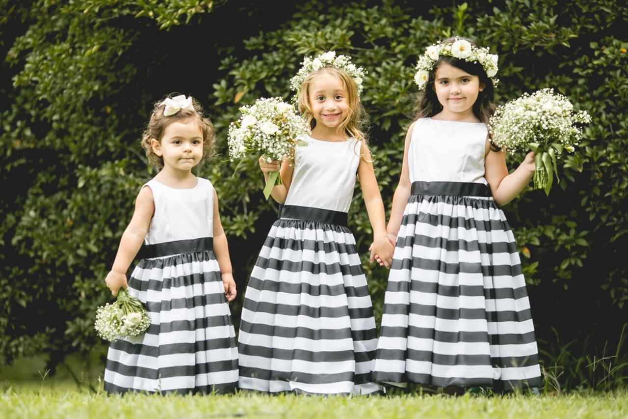 Três daminhas com vestidos listrados em preto e branco, com tiaras de flores na cabeça e mini buquês nas mãos