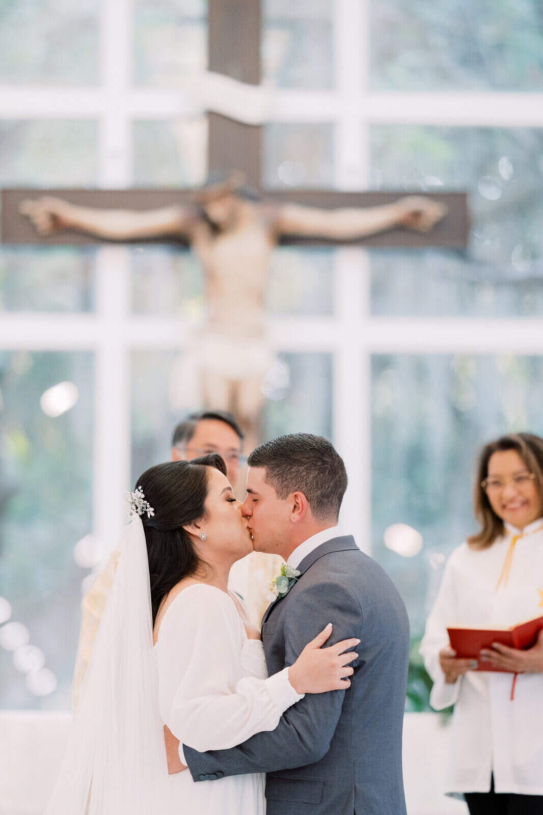 Casal de noivos se beijando em frente ao altar, com um padre e uma cruz com Jesus crucificado ao fundo
