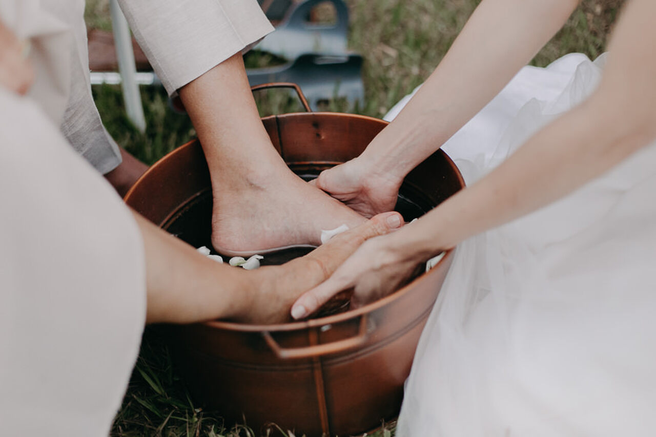 Pés do noivo dentro de uma bacia com água e mão da noiva sobre os pés dele lavando