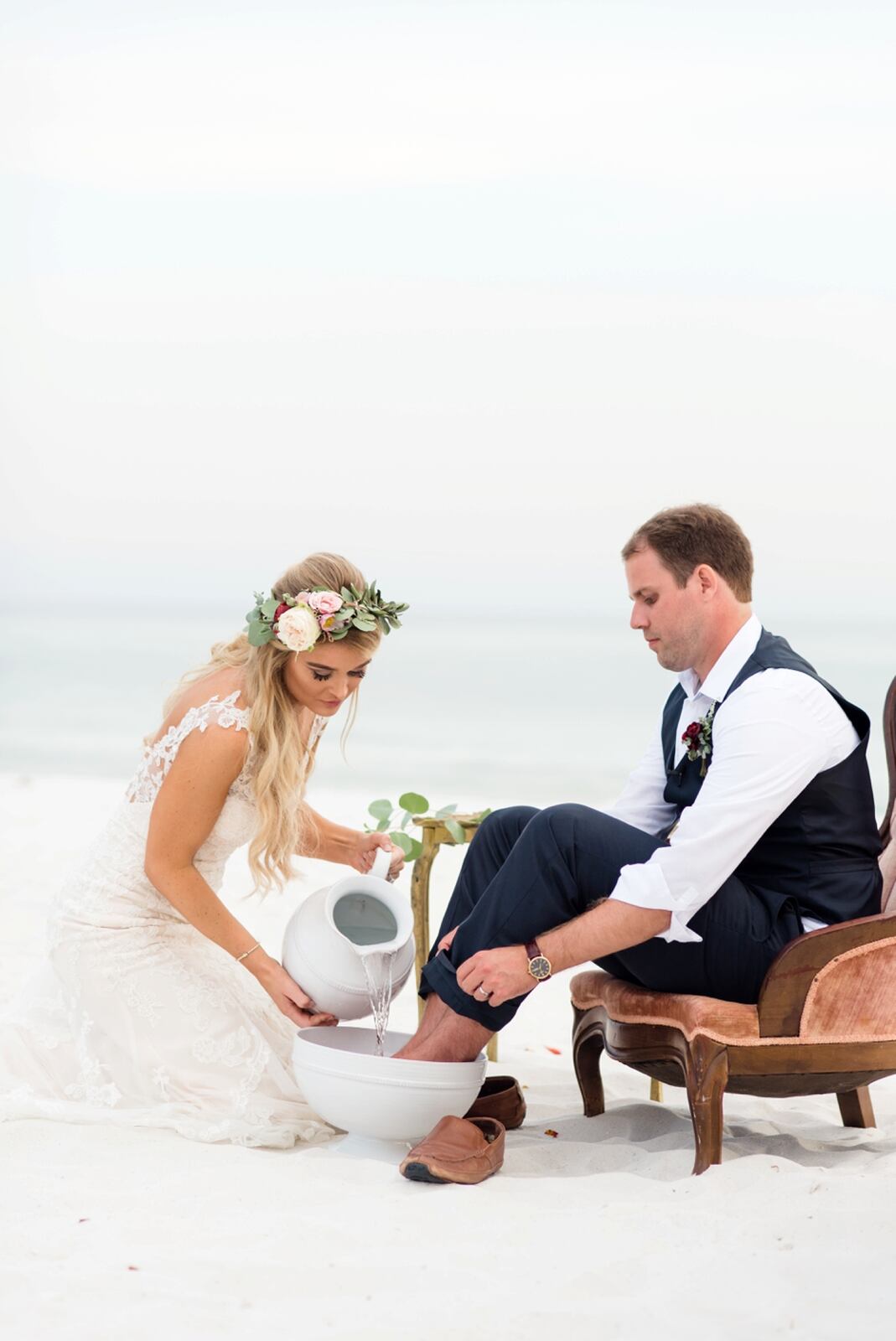 Noiva de joelhos com um jarro branco de água nas mãos derramando a água em cima dos pés do noivo que está sentado em uma cadeira à sua frente e com os pés dentro de uma bacia