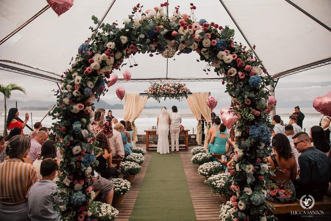 Cerimônia na praia, em um local coberto. Com um arco de flores no primeiro plano e ao fundo o casal de noivo no altar