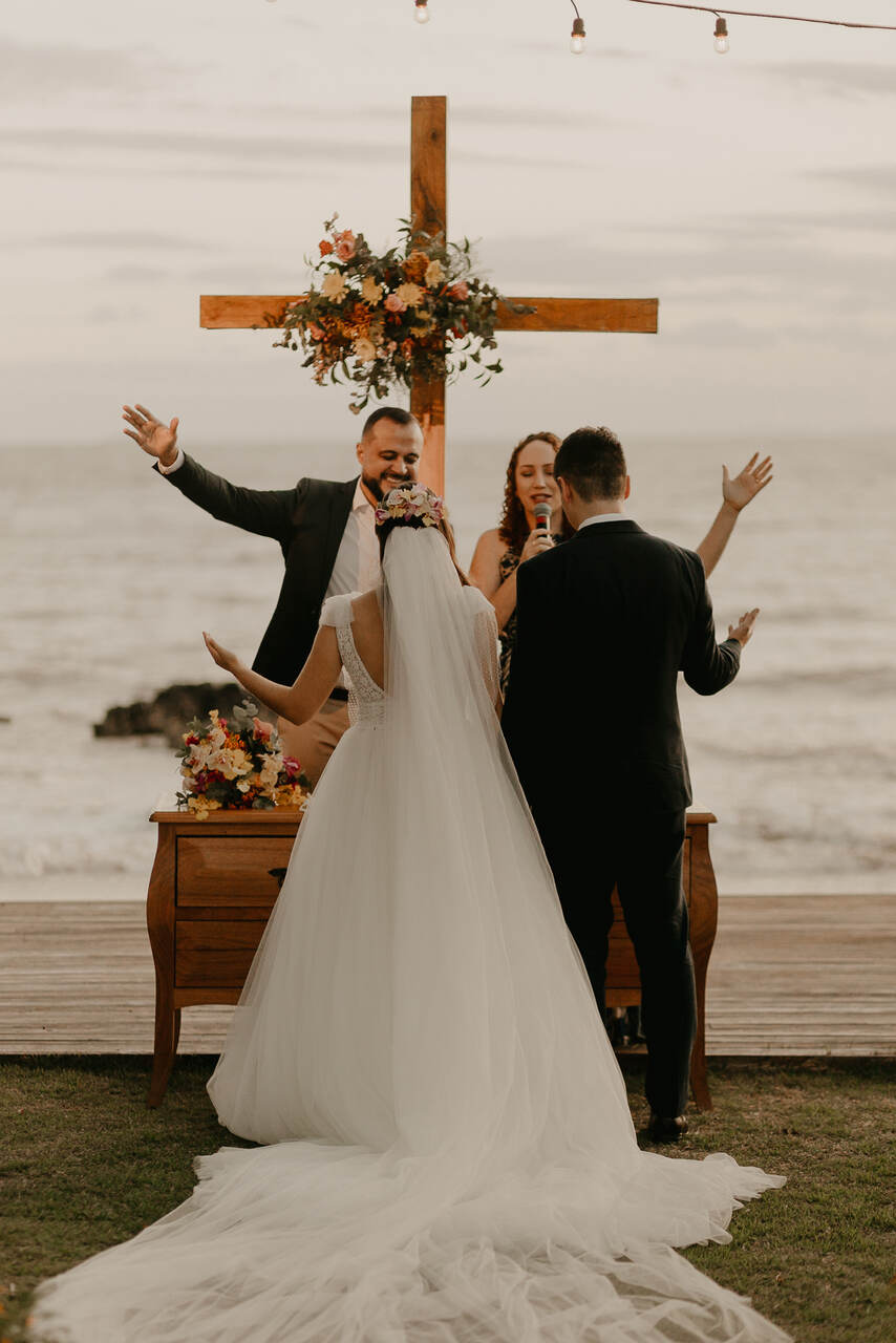 Casamento na praia. No altar os noivos estão com as mão levantadas enquanto os pastores oram