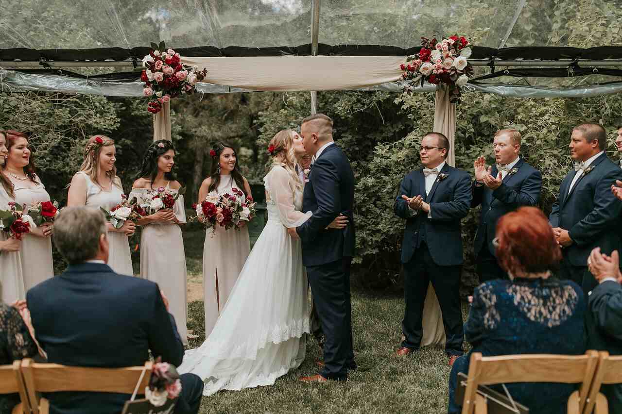 Noivos no altar se beijando, ao lado estão as damas e os bests man e o oficiante da cerimônia. No altar há um arranjos pequenos de rosas pendurados