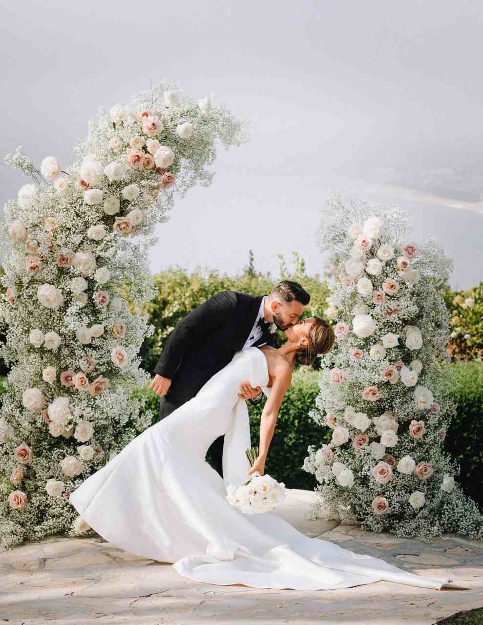 Casal se beijando no altar. Noivo se inclina para beijar a noiva que está segurando um buquê. No altar há um semi arco com flores mosquitinho e rosas 