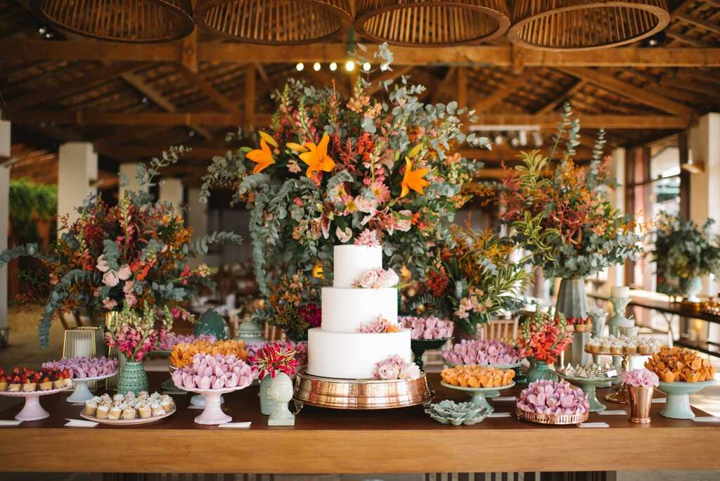 mesa do bolo de casamento com bandejas com doces e arranjos com flores vermelhas e laranjas