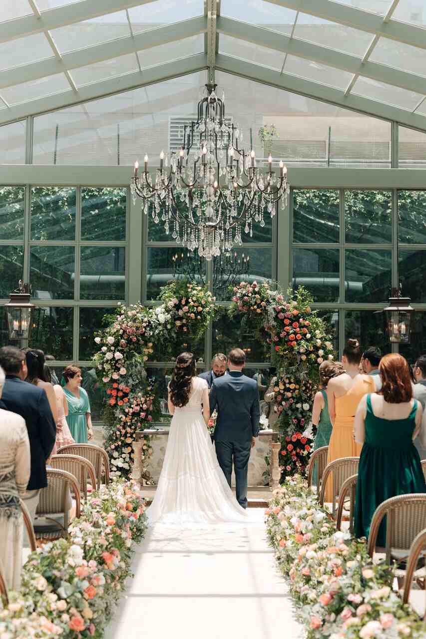 Noivos no altar, dentro de uma estufa de jardim. No altar há os noivos e oficial do casamento, um arco de flores e um lustre pendurado no teto.