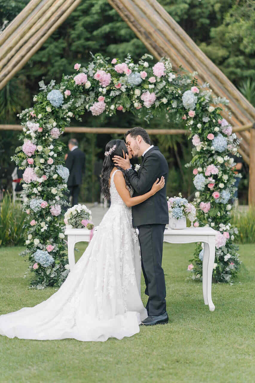 Casal se beijando no altar, com uma coroa de flores ao fundo e um aparador branco
