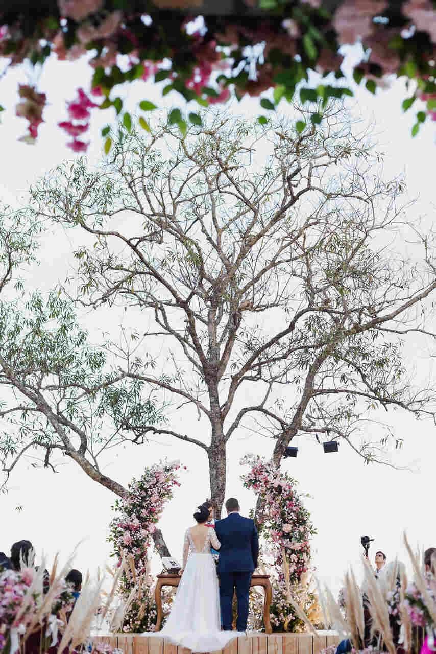 Cerimônia de casamento embaixo de uma árvore, com convidados assentados e os noivos no altar em frente a um arco de flores
