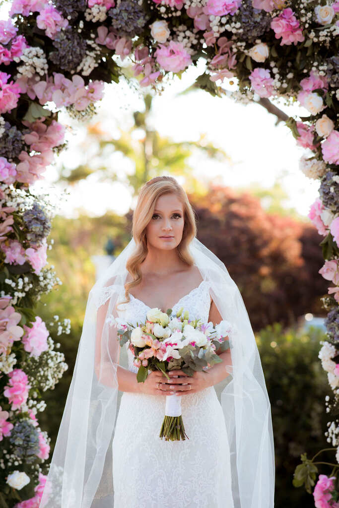 Noiva, posando para a foto, com um buquê de flores nas mãos e ao fundo um arco de flores