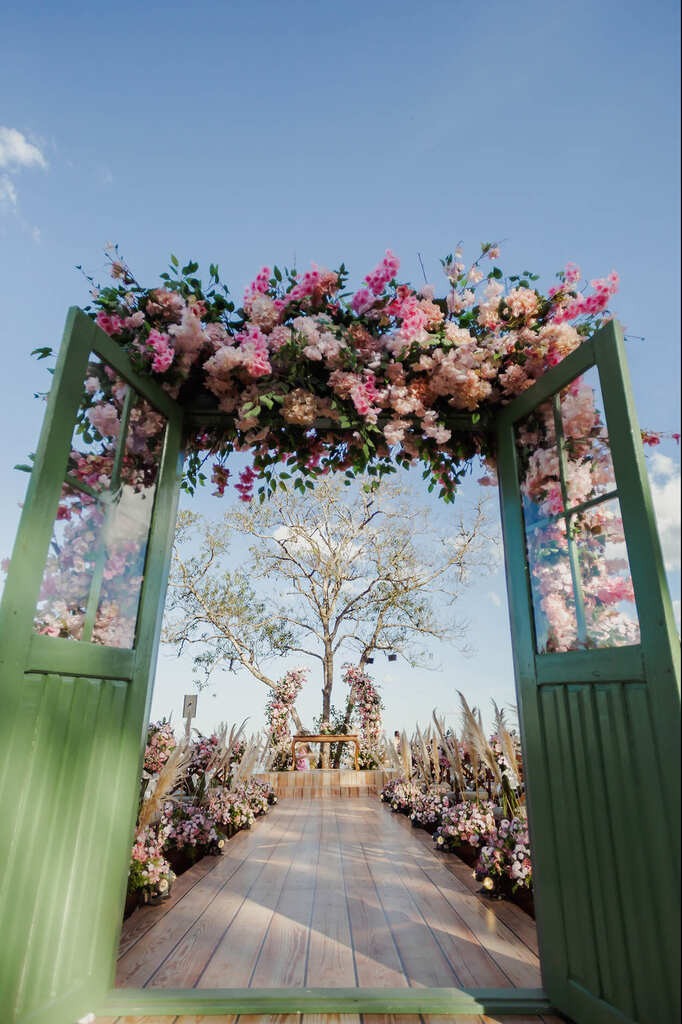 Porta de madeira na entrada da cerimônia, com flores em cima da porta. Ao fundo, no final da passadeira, está o altar com um arco de flores